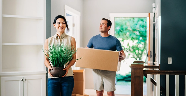 Woman carrying plant and man carrying cardboard box into new home with freshly painted walls.