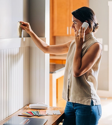 Woman speaking on cell phone holding paint swatch up to wall.