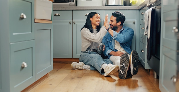 A couple sitting on a kitchen floor giving each other a high five.