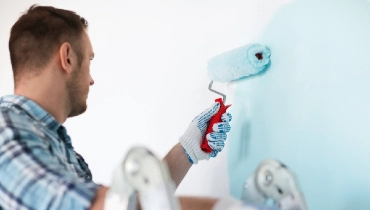 Man wearing work gloves and using a roller to paint an interior wall.