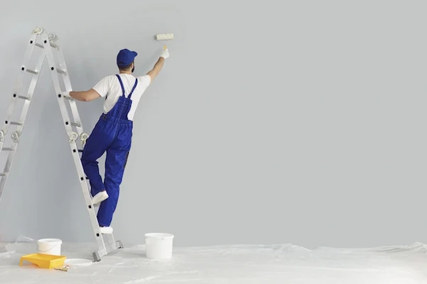 Male painting technician in blue overalls holding paint roller and standing on ladder.